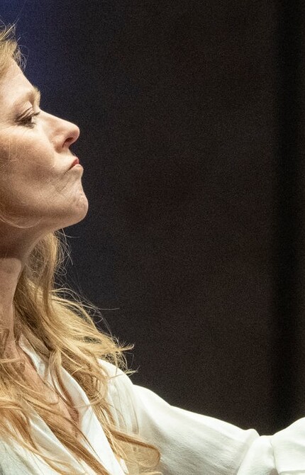 A person with long blonde hair, wearing a white blouse, conducting with an intense expression.