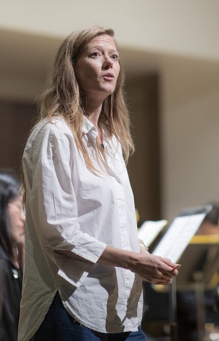 Person in a white shirt standing on stage, surrounded by an ensemble in performance attire, speaking to the audience, creating a connection before or during the performance.