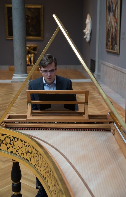 A person wearing a blue suit and glasses plays an antique grand piano in a museum setting.