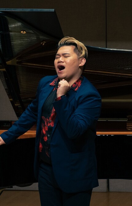 Person in a blue suit and black shirt with red flowers singing on stage, with one hand resting on a grand piano, exuding confidence and emotion in the performance.