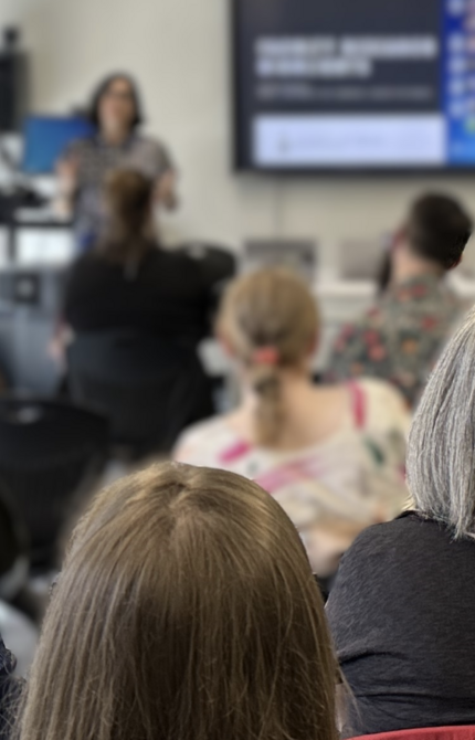 Group of people sitting in a classroom, attentively listening to a workshop, focusing on the speaker at the front.