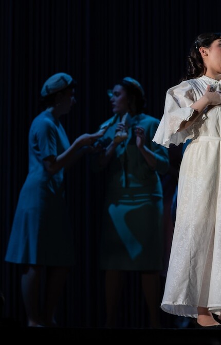 Scene from an opera, with a person singing in a white dress, while others are conversing in the background.