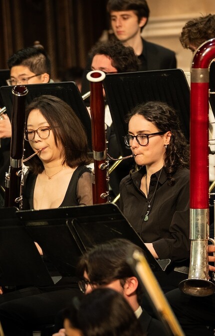 Four bassoon players in performance attire playing together as part of an ensemble, each focused on their music stands.