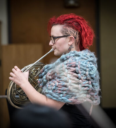 A person with red hair playing the French horn, viewed from the side, wearing a knit wrap.