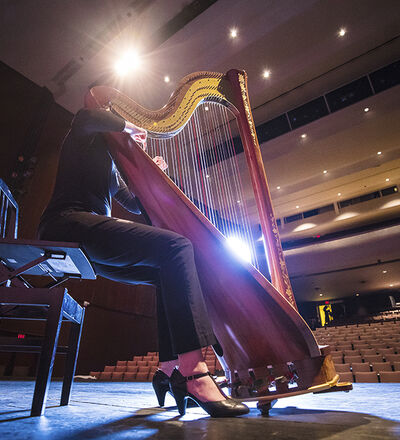 Person in performance attire playing the harp on stage, performing for an empty auditorium, with rows of seats and stage lighting in the background.