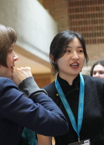 A person in a black dress giving a presentation to two interested individuals, pointing to an information board in the Edward Johnson Building lobby.