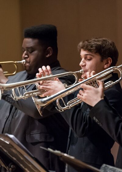Four students playing trumpets on stage as part of an ensemble performance, dressed in concert attire and illuminated by stage lighting.