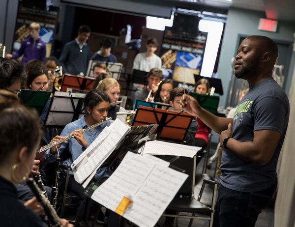 Man conducting an ensemble in a classroom, leading with a baton while the musicians are focused on him.