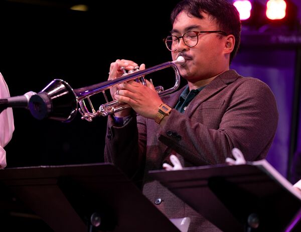 Person wearing a suit playing a muted trumpet on stage with a music stand in front of them