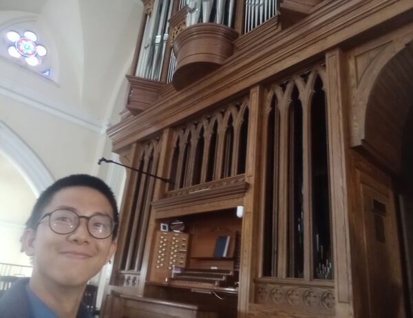 Person with glasses smiling while standing in front of a large organ, showcasing the instrument's intricate pipes and design.