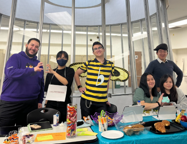 Griffin Hook is shown wearing a bumble bee costume with the musical symbol of a flat on his shirt (it is a "pun costume"; he is a B flat.) He is surrounded by other U of T Music students, and they are holding a bake sale.