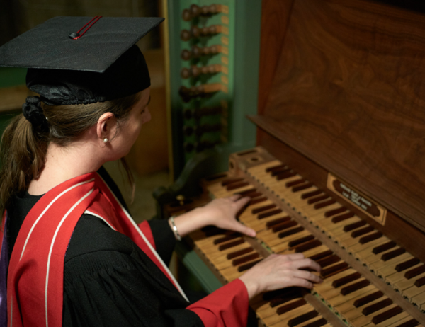 Person playing the organ while wearing a graduation cap and gown, focused on the keyboard.
