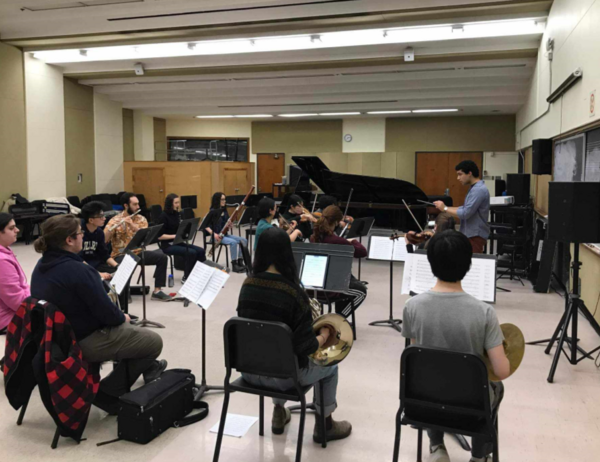 Music students in a classroom playing in an ensemble, conducted by a person in a grey shirt waving a conductor's wand, guiding the group through the performance.
