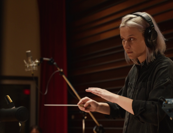 Person wearing headphones and a black shirt conducting in a music studio, surrounded by microphones, with wood panels and a red curtain in the background