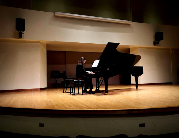 Person playing grand piano on the Walter Hall stage, with stage lighting.