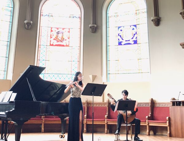 Person in a dress standing and playing the flute, while another person sitting plays the guitar, in a chapel setting.