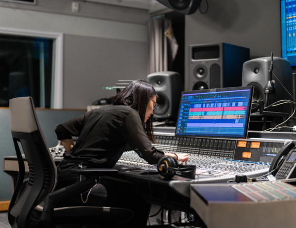 Person wearing all black working in an electronic music studio, surrounded by equipment like synthesizers, mixers, and soundboards, focused on their work.