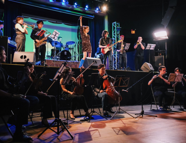 Ensemble performing on stage with a vocalist, captured from the audience perspective, showcasing the musicians and singer in action during the performance.