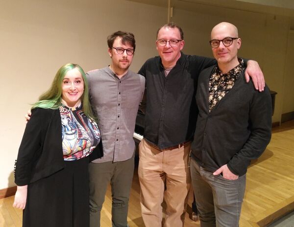 Group photo of four people standing together, with wood floors beneath them and a white wall in the background, creating a simple and clean setting.