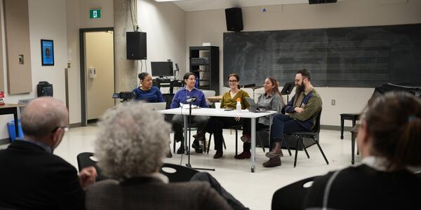 Panel discussion in a classroom, with a group of people watching as five individuals speak around a table, engaging in conversation and sharing insights.