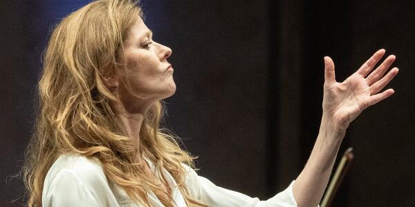 A person with long blonde hair, wearing a white blouse, conducting with an intense expression.