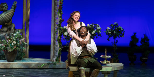 Two performers smiling on stage in a garden scene.