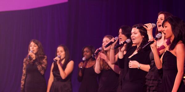 Choir performing wearing all black, in front of a black background with purple lights illuminating the scene.