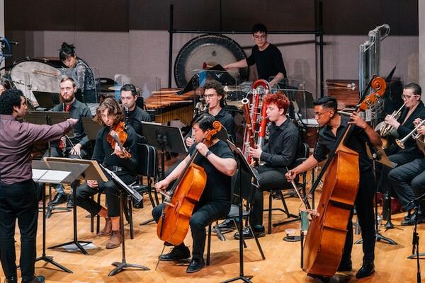 Ensemble on stage wearing performance attire, seated and waiting attentively to be cued by a conductor in a burgundy shirt.