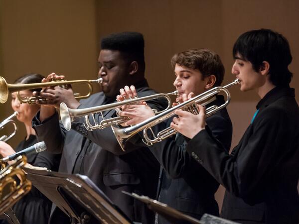 Four students playing trumpets on stage as part of an ensemble performance, dressed in concert attire and illuminated by stage lighting.