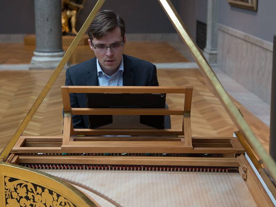 A person wearing a blue suit and glasses plays an antique grand piano in a museum setting.