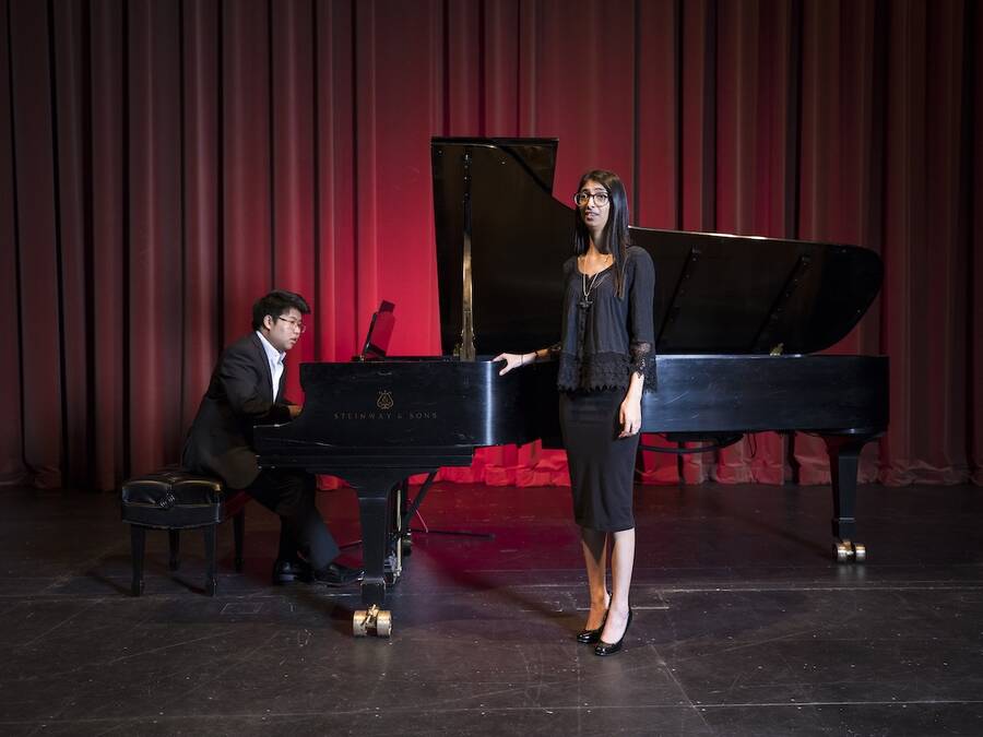 Vocalist and accompanist performing on stage, with the vocalist singing and the accompanist playing a grand piano, set against a red curtain backdrop.