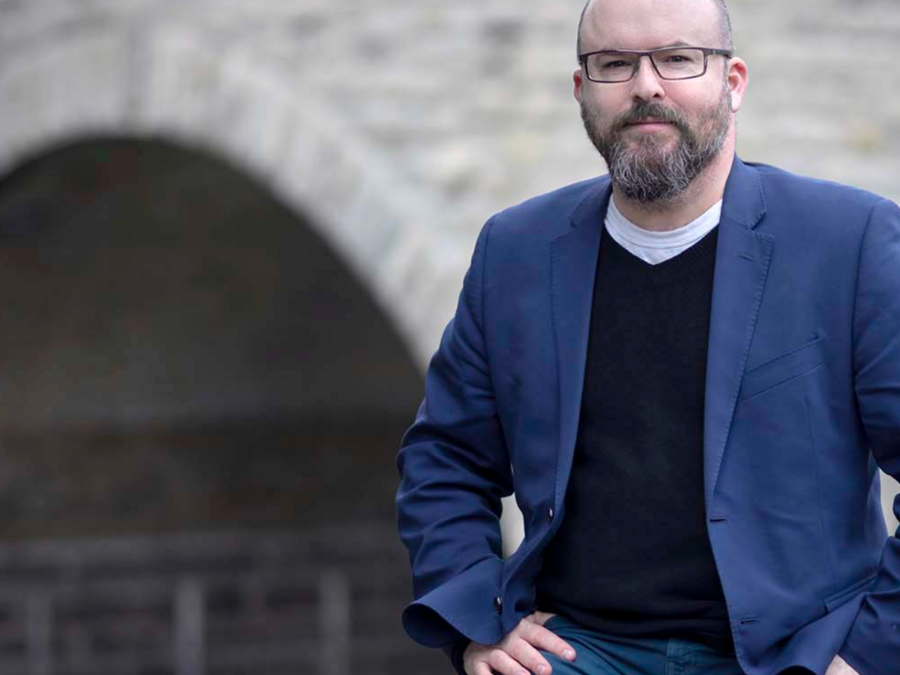 A person with glasses is wearing a black sweater, navy blazer, and jeans, posed in front of an old stone building.