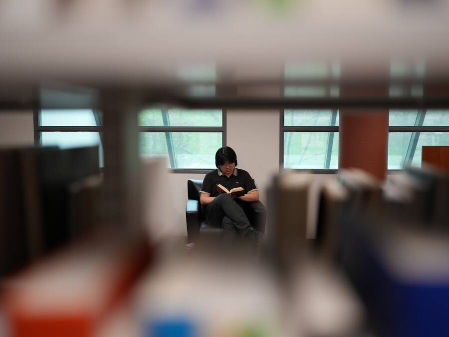 Photo of a person reading a book, with the camera positioned to capture the shot through a shelf of books in a library.