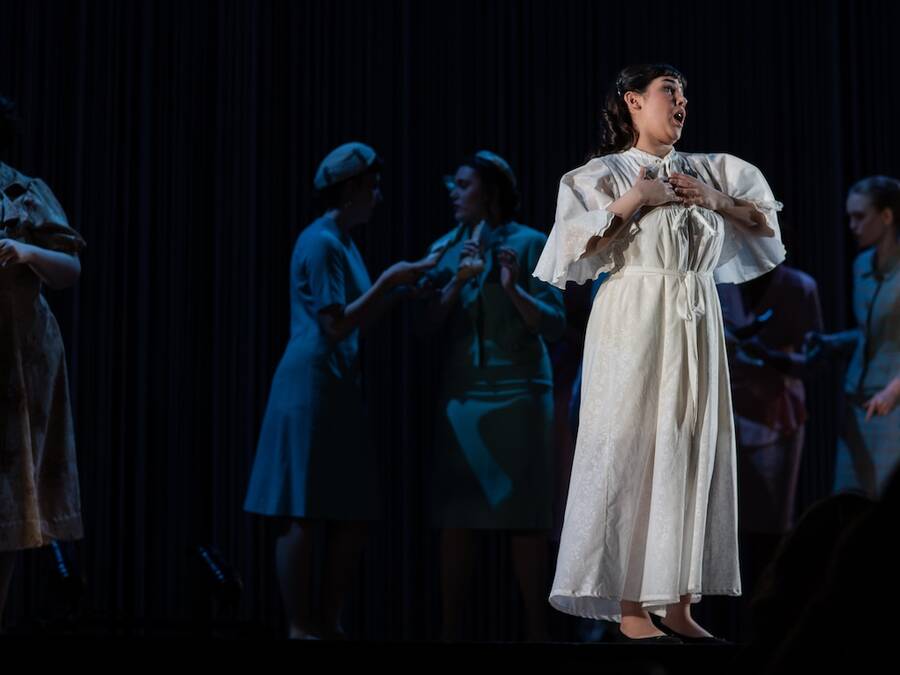 Scene from an opera, with a person singing in a white dress, while others are conversing in the background.