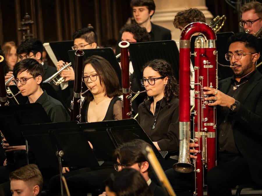 Four bassoon players in performance attire playing together as part of an ensemble, each focused on their music stands.