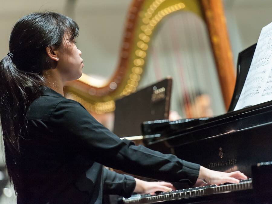 Person with hair in a ponytail, wearing performance attire, playing the piano in an ensemble, with a harp visible in the background.