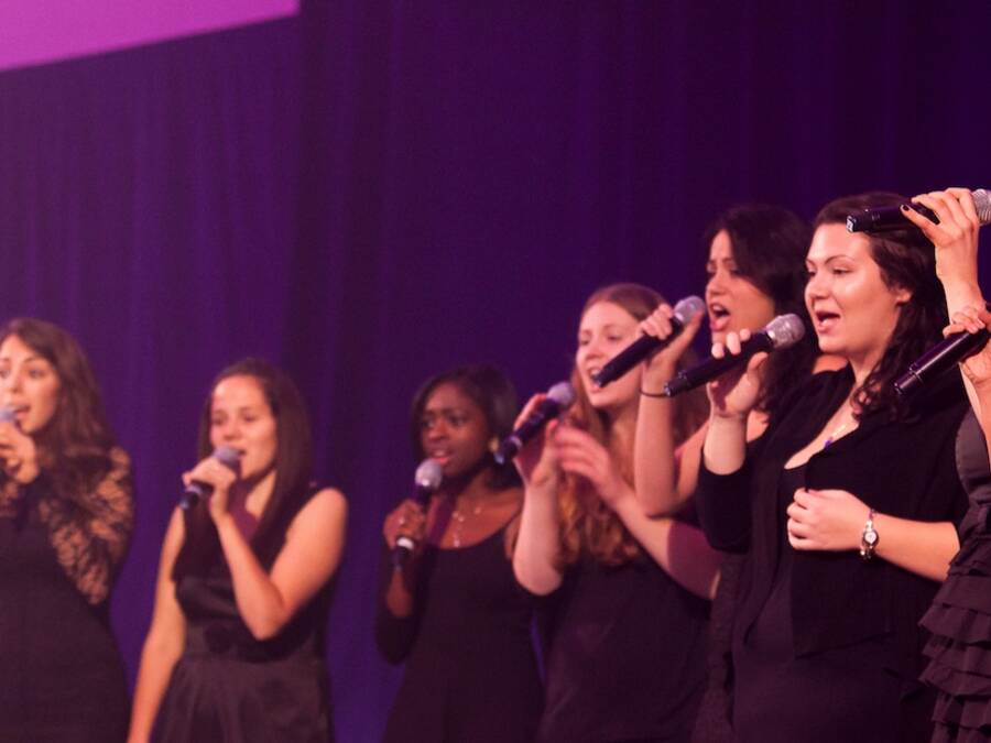 Choir performing wearing all black, in front of a black background with purple lights illuminating the scene.