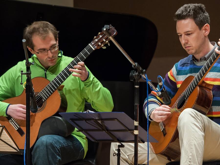 Two guitarists performing, surrounded by performance equipment, both focused on the music on a stand.