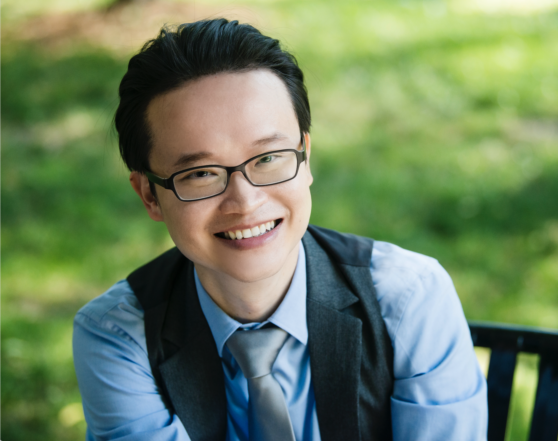 Professional headshot of Charles Lin, smiling and sitting on a bench with greenery behind him.