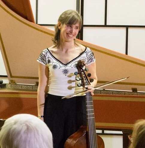 Picture of a woman standing in front of a piano, smiling and looking to the side while holding her instrument, wearing a white top and black pants.