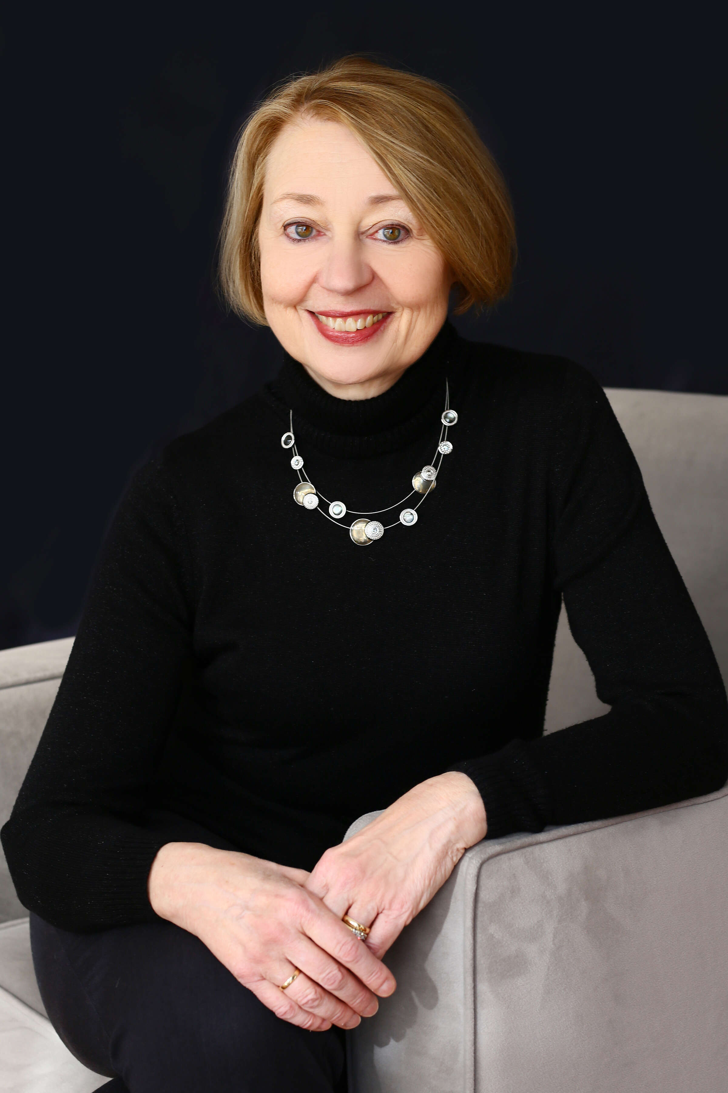 Photo of a woman wearing all black and a silver necklace, posed in a grey chair.