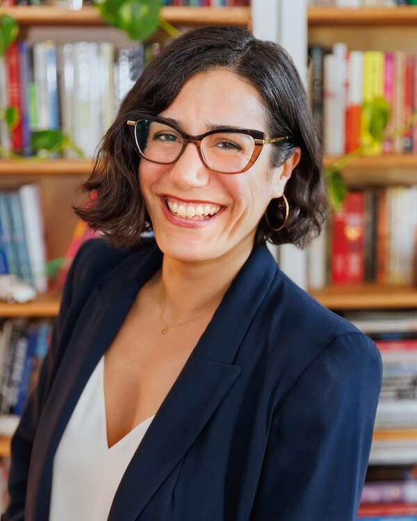Headshot of a person smiling, wearing glasses and a blue blazer, with bookshelves in the background.
