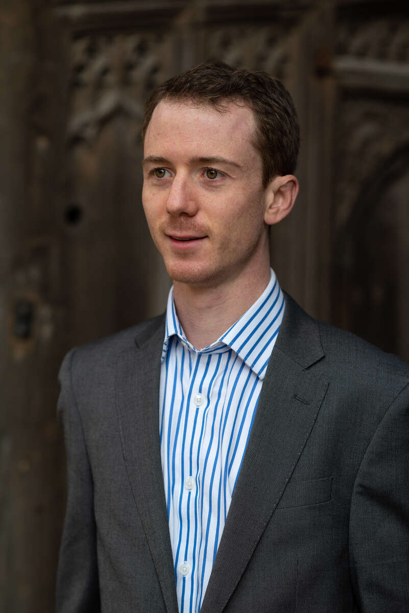 Person with short hair, wearing a shirt and blazer, standing in front of a stone wall with carvings.