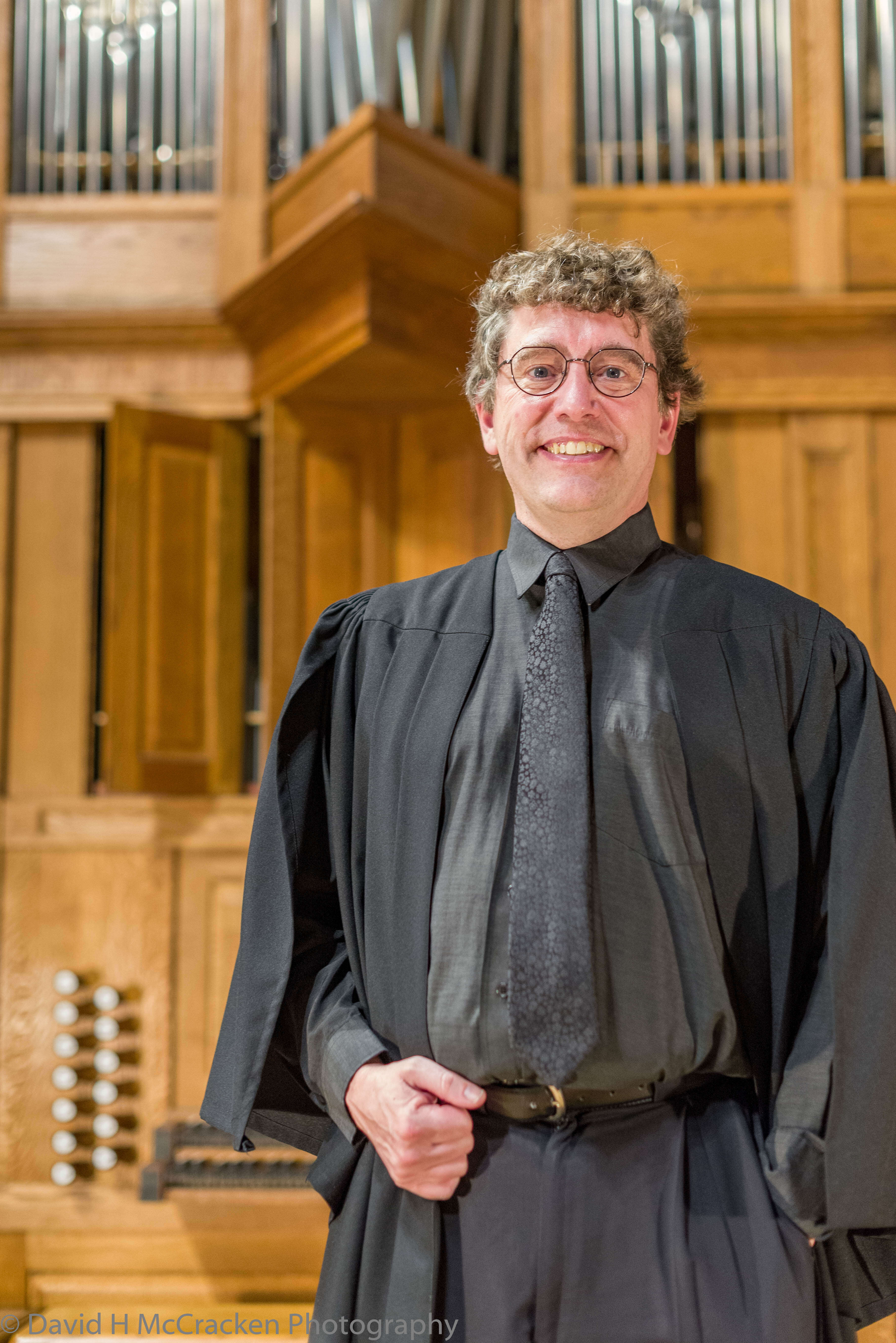 Person wearing glasses and an organist robe, standing in front of an organ.