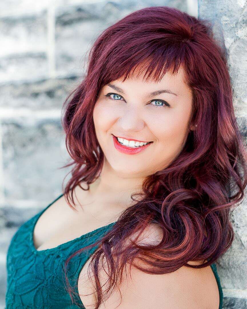 Photo of a person with long red hair, wearing a blue tank top, standing in front of a brick wall.