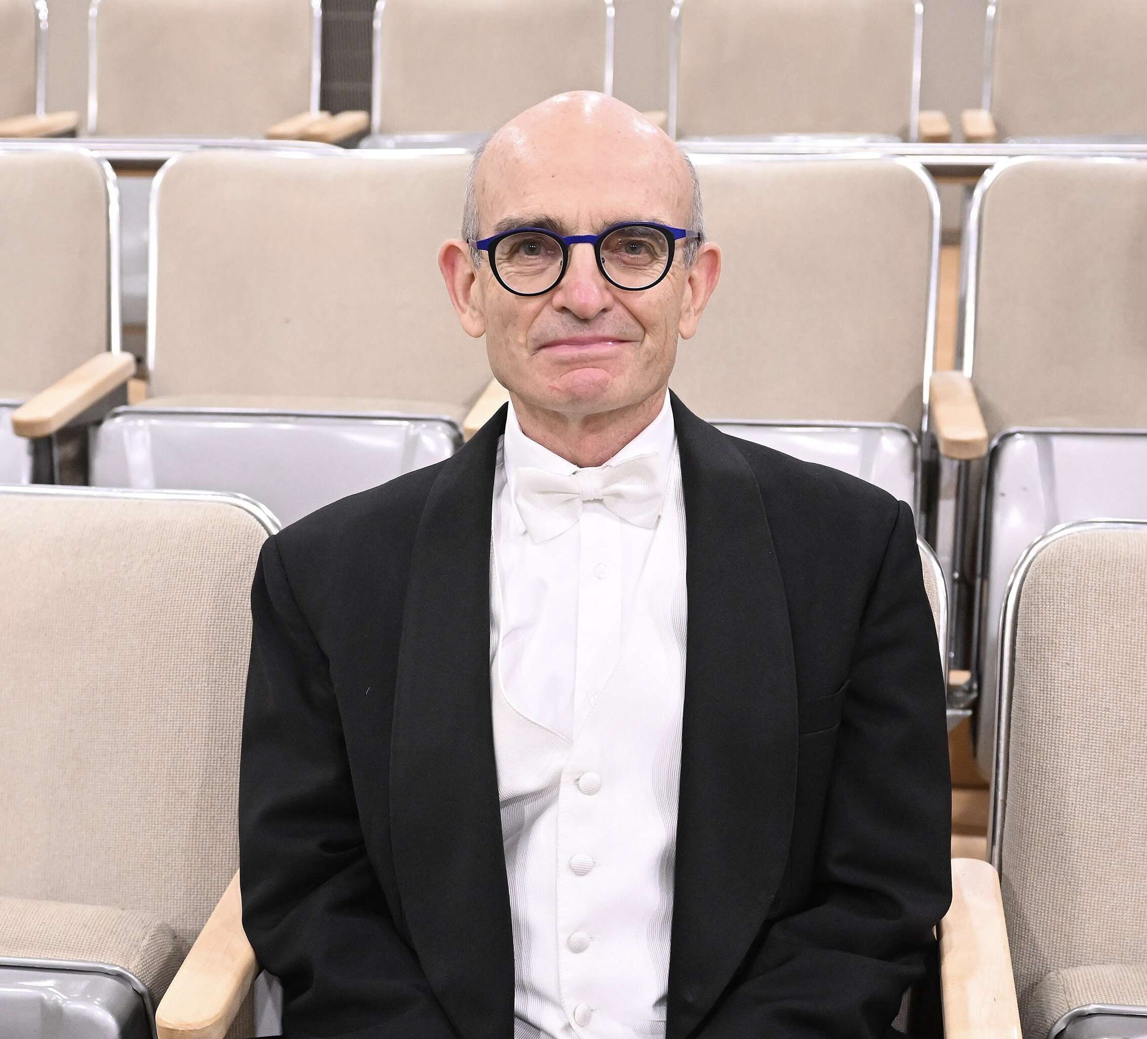 Close-up picture of John Rudolph sitting in the middle of a row of seats with a professional suit. 