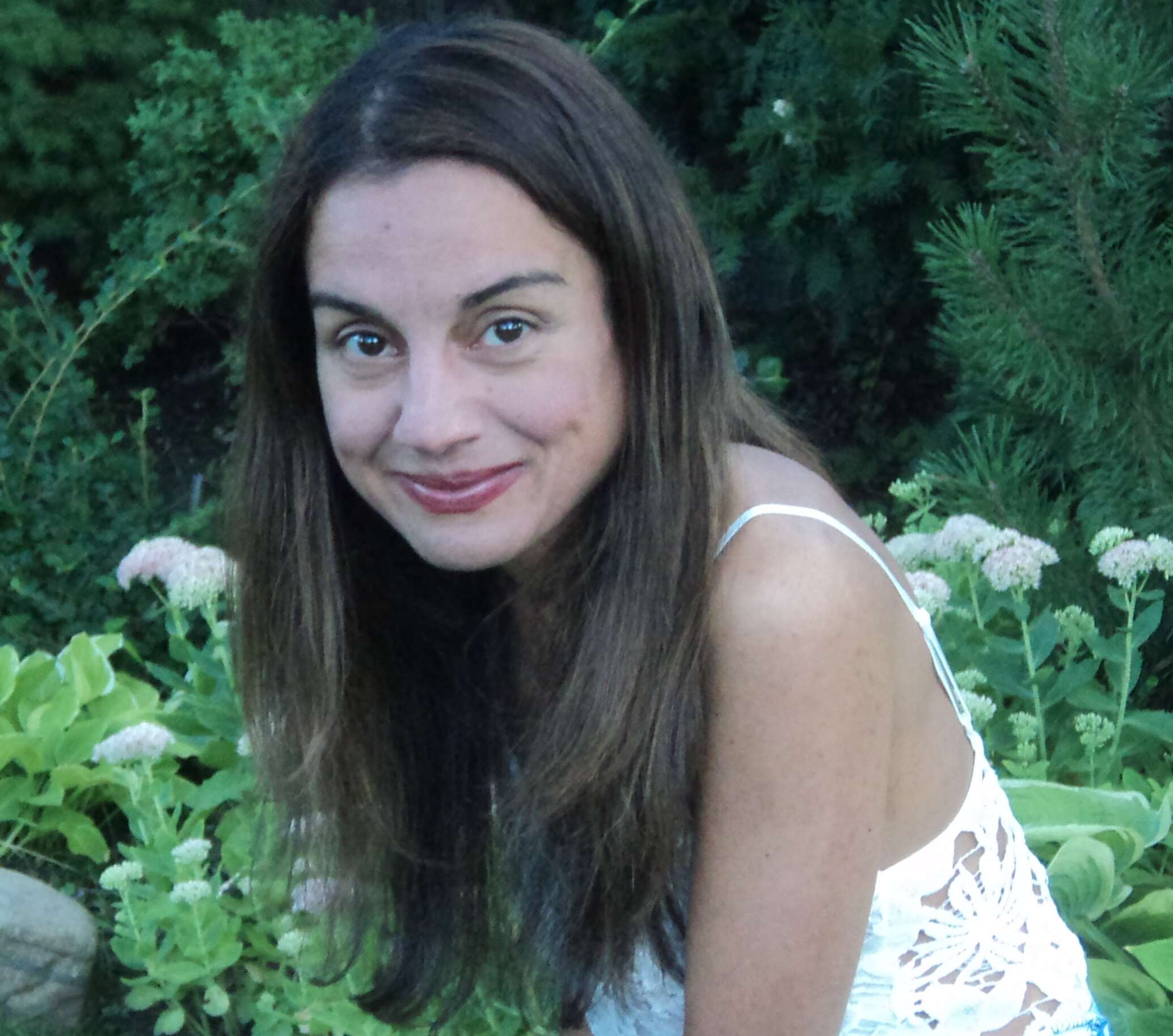Smiling woman with long hair wearing a white dress, sitting in a garden surrounded by plants and flowers.
