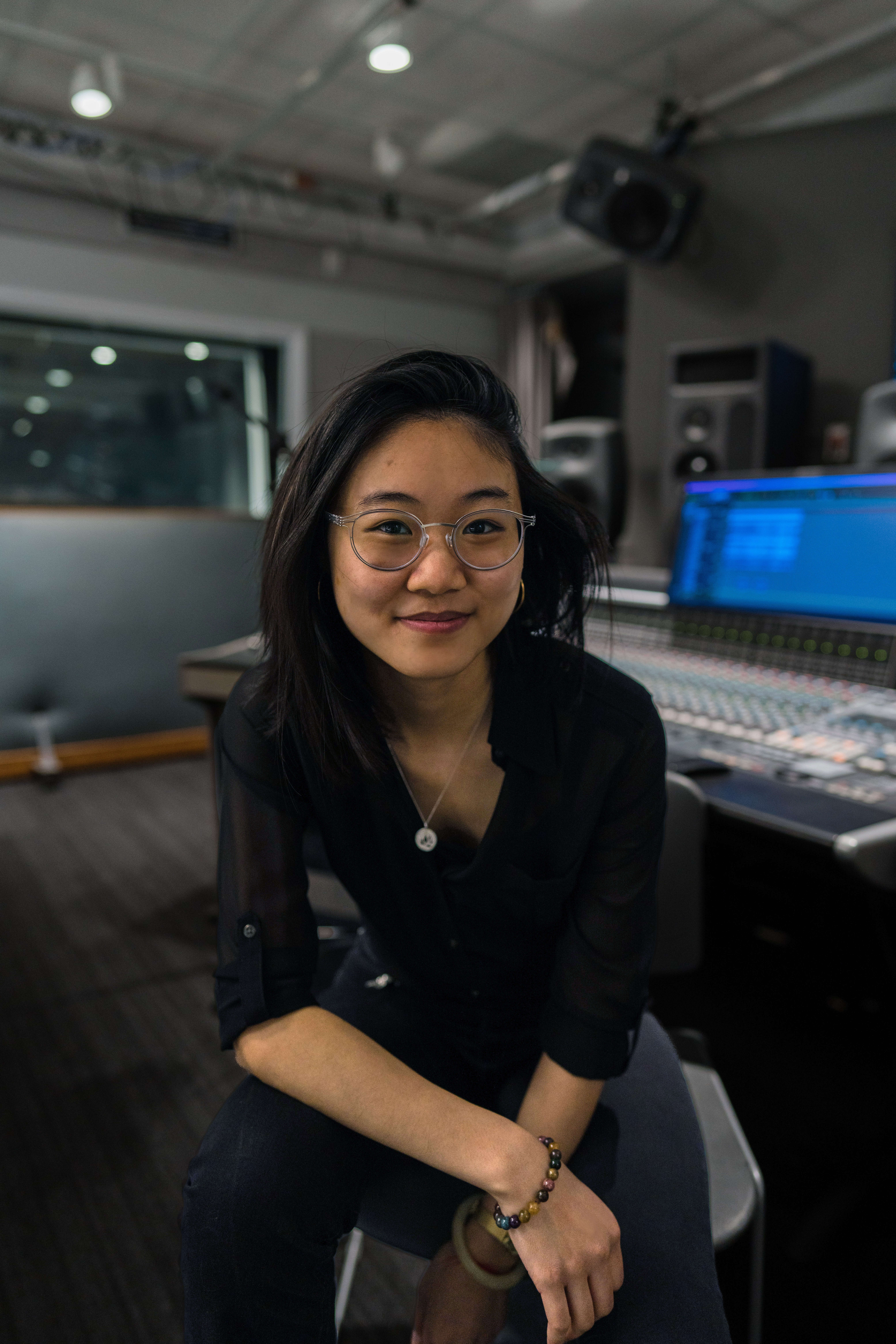 Person wearing a black button-down shirt sitting on a stool in the electronic music studio, surrounded by equipment and sound gear.
