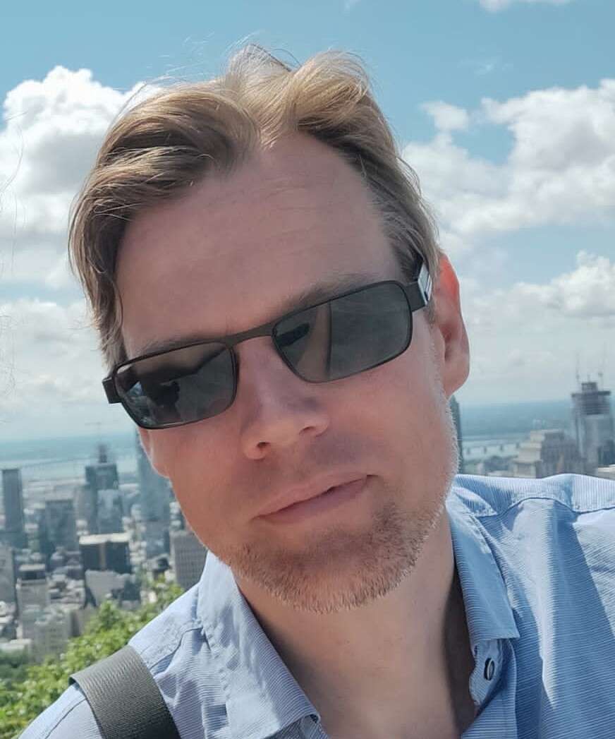 Photo of a man wearing sunglasses and a blue collared shirt, standing in front of a city skyline.
