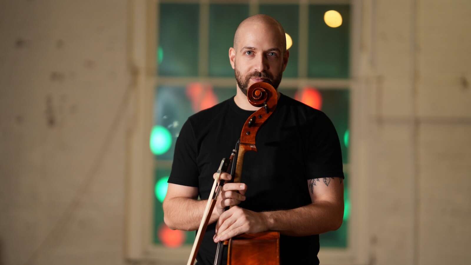 A bald person wearing a black t-shirt, holding a cello, in front of a wall with a window with blurred lights in the background.